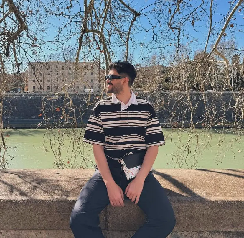 Kelvin Maciel, um homem, sentado em mureta de pedra diante de um rio. Veste camisa polo listrada e óculos escuros. Galhos de árvores emolduram a paisagem urbana ao fundo em dia ensolarado.