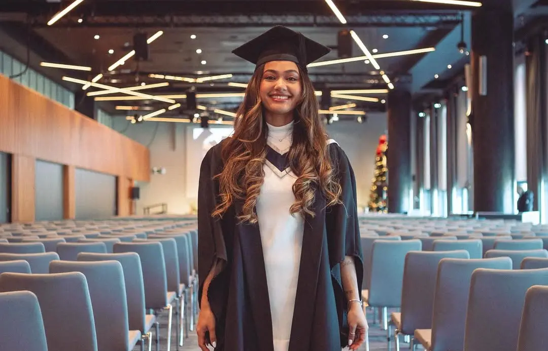 Jovem sorridente em traje de formatura, beca e capelo, no centro de um grande auditório com fileiras de cadeiras vazias. Luzes lineares no teto e árvore de Natal ao fundo.