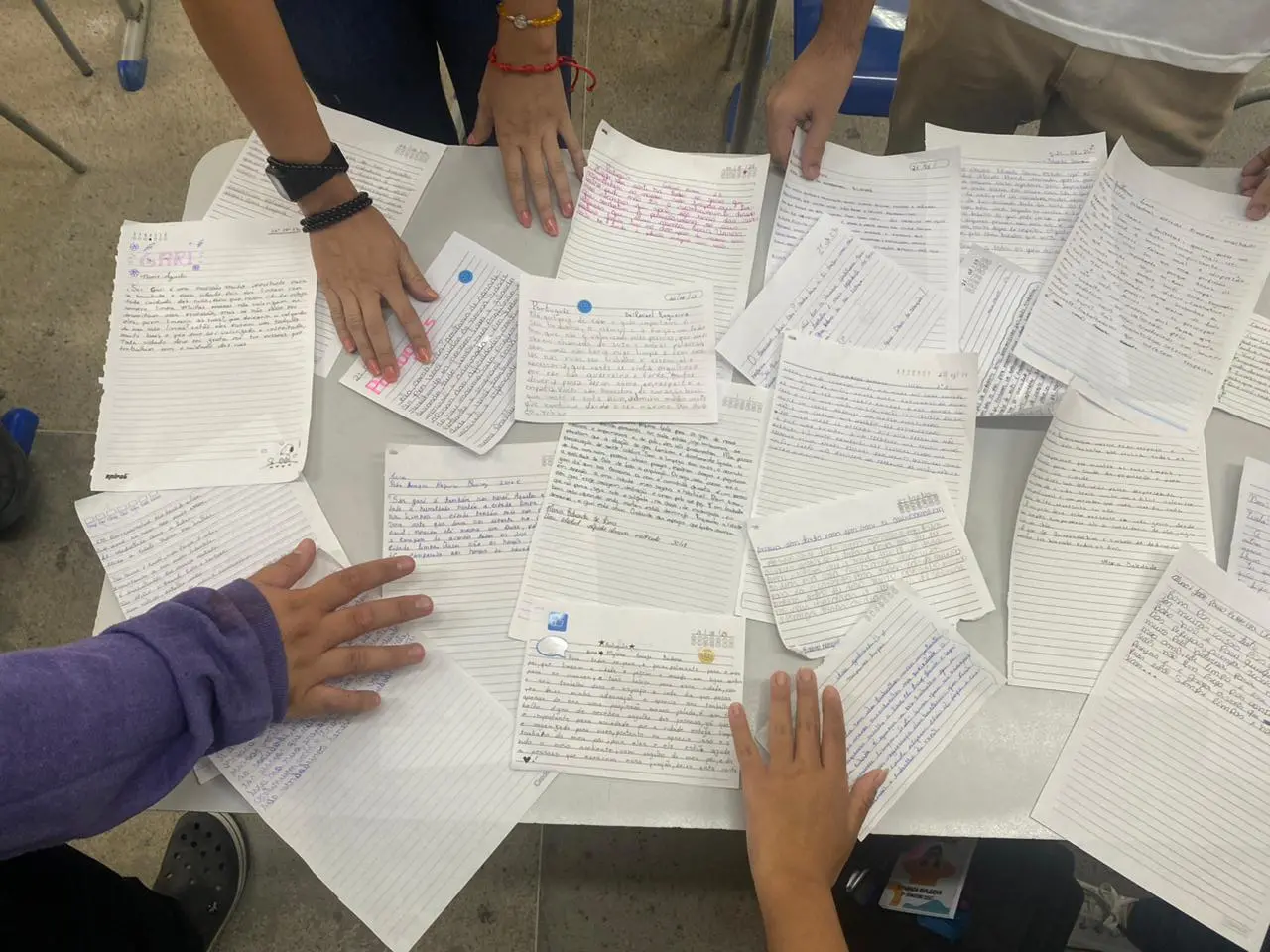 Na imagem, vista superior de uma mesa branca onde diversas mãos de diferentes pessoas tocam e organizam várias folhas de papel espalhadas. As folhas contêm textos escritos à mão, alguns com canetas de cores diferentes (azul, preto e rosa) e pequenos adesivos ou desenhos. Os papéis parecem ser cartas ou mensagens de agradecimento e relatos. As mãos ao redor da mesa sugerem um momento de compartilhamento ou revisão coletiva do material produzido.