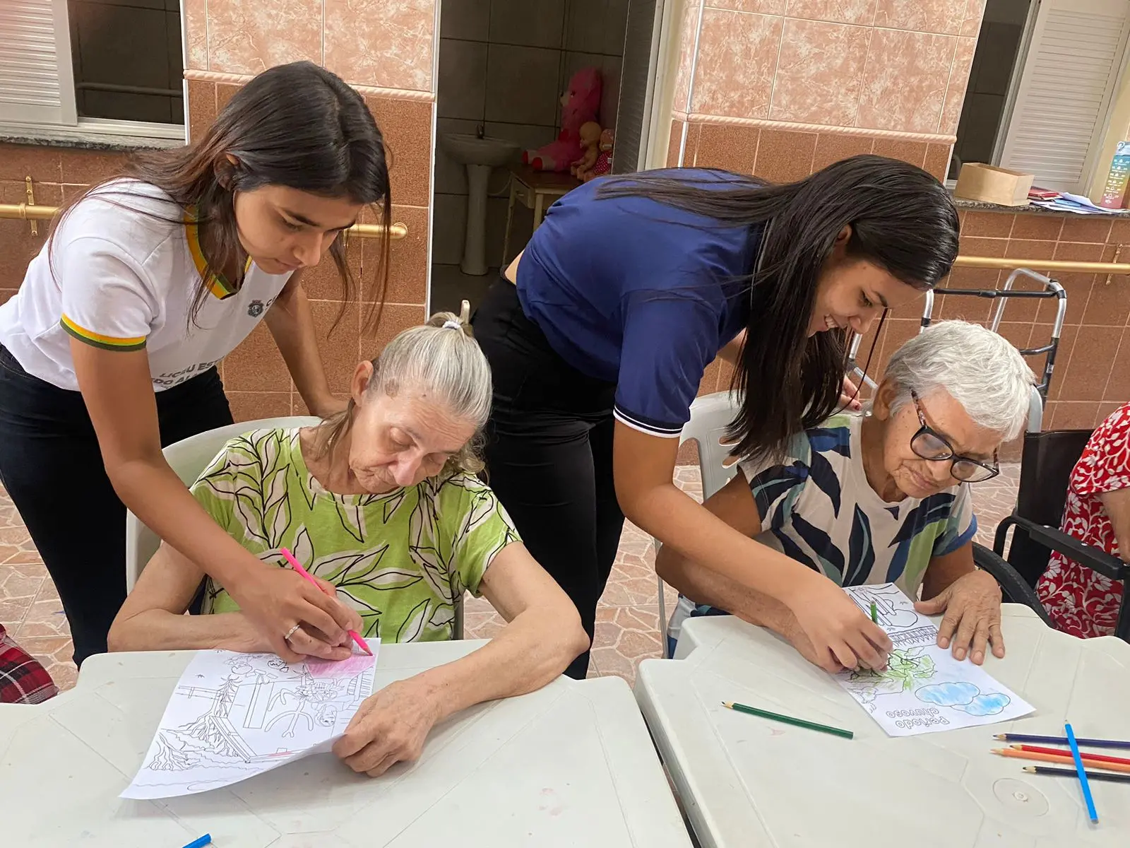 Na imagem, duas jovens estão inclinadas sobre uma mesa branca, auxiliando duas senhoras idosas em uma atividade de pintura. A jovem à esquerda usa um uniforme escolar branco e orienta a senhora à sua frente, que pinta um desenho com um lápis cor-de-rosa. À direita, a outra jovem, de camiseta azul marinho, observa sorridente enquanto a segunda senhora colore um desenho de paisagem com lápis verde. Sobre a mesa, há diversos lápis de cor espalhados. O ambiente é iluminado e possui paredes revestidas com azulejos em tons de bege.