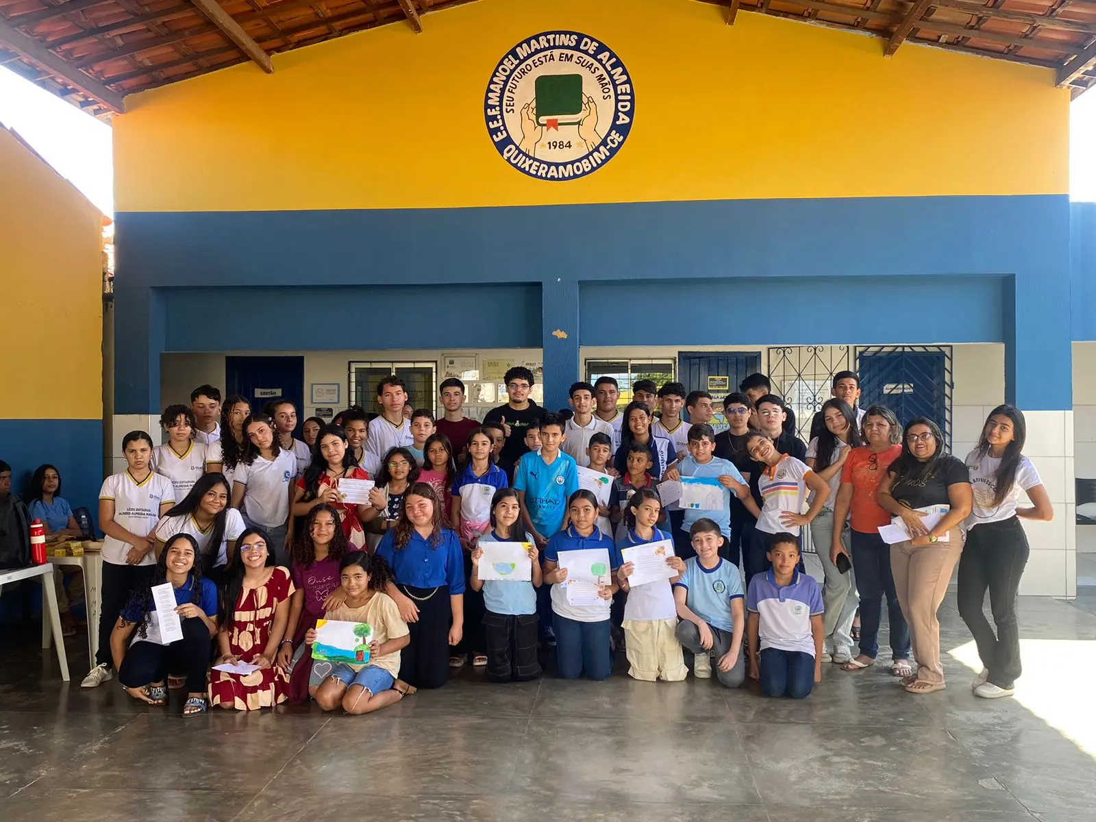 Na imagem, um grande grupo de aproximadamente quarenta pessoas, majoritariamente crianças e adolescentes, posa para uma foto coletiva em um pátio escolar coberto. Eles estão posicionados à frente de uma parede pintada de amarelo e azul, que exibe no topo o brasão da 