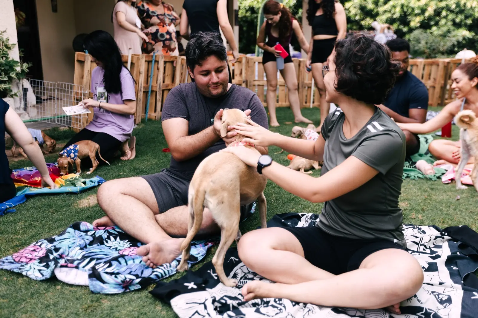 Casal sentado na grama acaricia filhote de cachorro caramelo em evento de ioga e adoção. Ao fundo, outros participantes e um cercado de madeira.