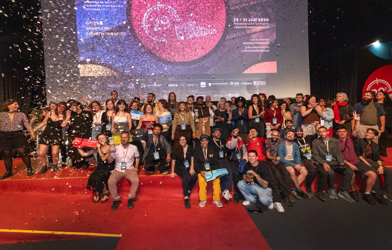 Um grupo numeroso de pessoas celebra no palco da 29ª Mostra de Tiradentes sob uma chuva de confetes prateados, com alguns segurando troféus e prêmios simbólicos. Ao fundo, um telão exibe a identidade visual do evento em tons de rosa e azul, completando a atmosfera festiva e vibrante da premiação.