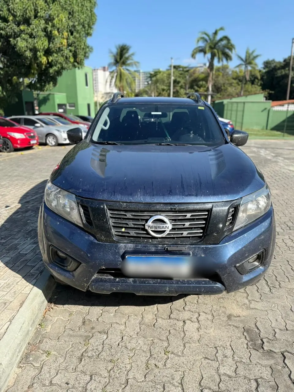 Vista frontal de uma picape Nissan azul escura estacionada em área externa, com sinais de poeira na lataria e placa borrada, em local que aparenta ser um pátio ou estacionamento.