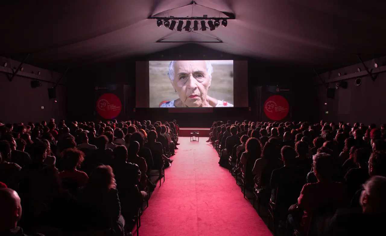 Uma plateia lotada ocupa o cinema escuro da 29ª Mostra de Tiradentes. Na tela, brilha o rosto em close de uma mulher idosa. Um corredor de tapete vermelho divide as poltronas e conduz o olhar até a projeção, em uma atmosfera solene de contemplação cinematográfica.