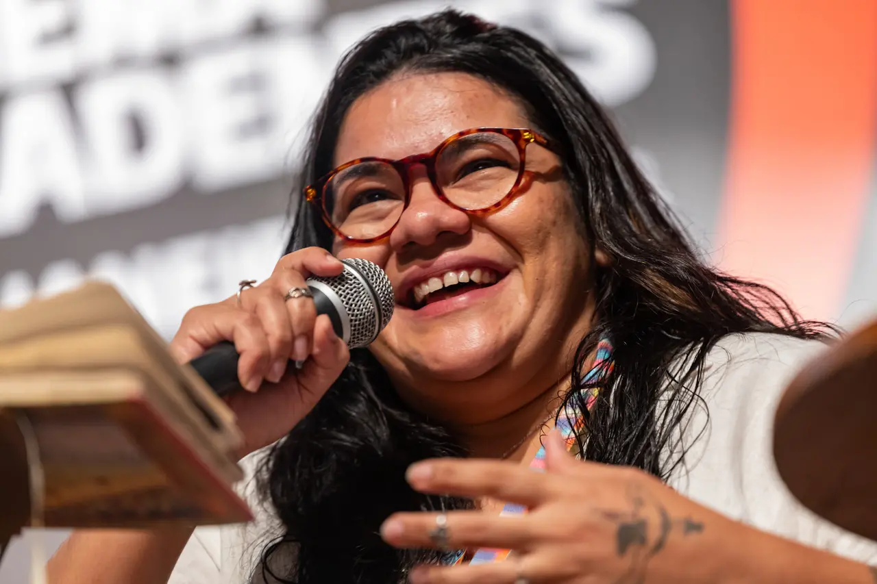 A diretora Priscila Smiths sorri entusiasticamente enquanto fala ao microfone durante um debate na 29ª Mostra de Tiradentes. Usando óculos de armação de tartaruga, ela gesticula com as mãos tatuadas em uma composição vibrante que destaca sua expressão de alegria sob o fundo desfocado do evento.