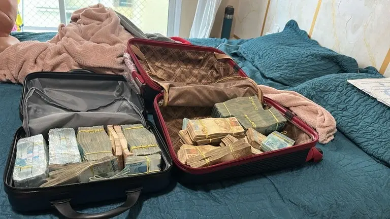 A imagem mostra duas malas abertas em cima de uma cama, com uma grande quantia de dinheiro em espécie.