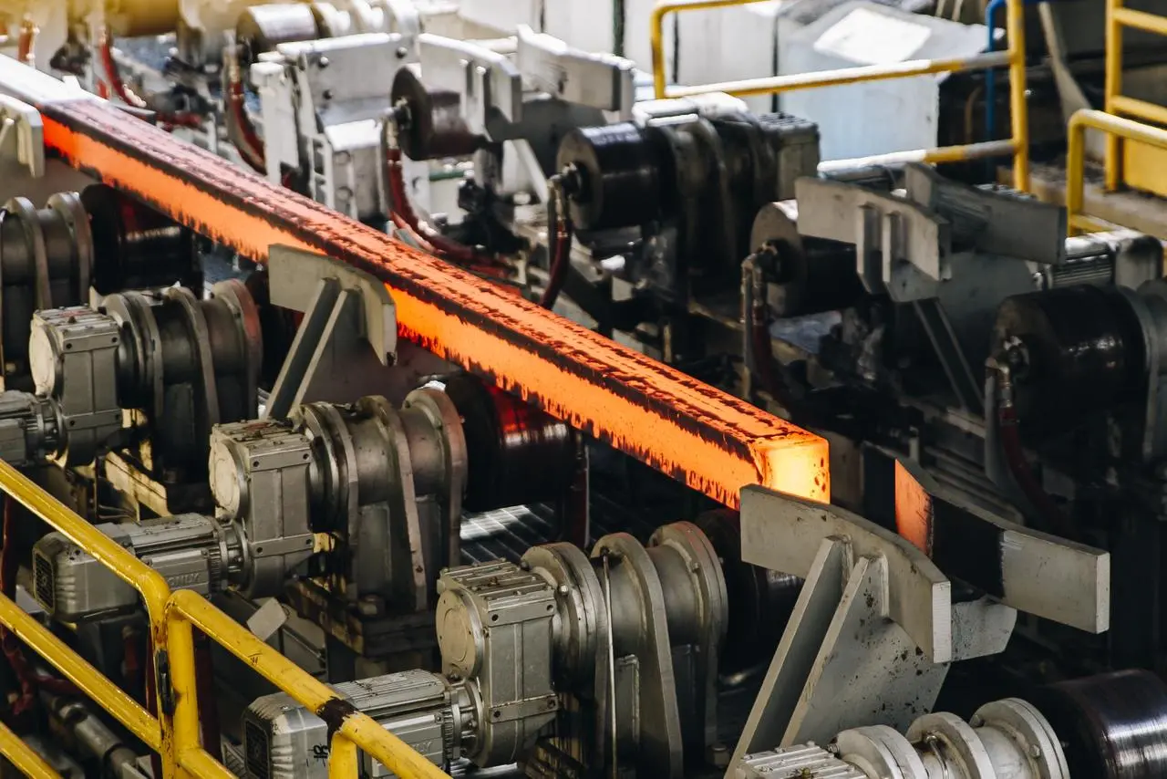 Barra de aço incandescente segue por rolos em linha industrial.