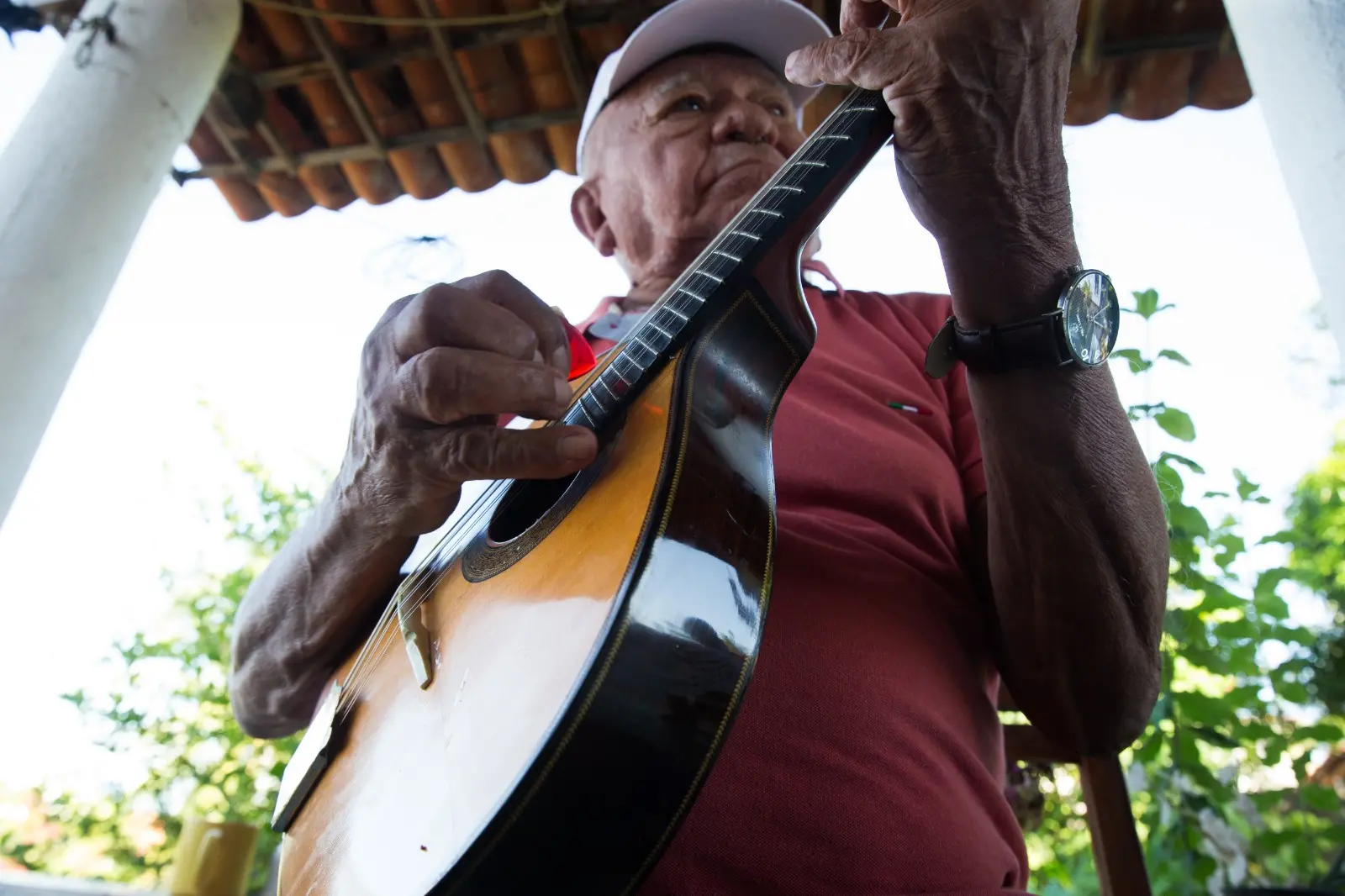 Na imagem, fotografia em ângulo de baixo para cima (contra-plongée) focando um homem idoso tocando bandolim. A perspectiva destaca o instrumento em primeiro plano, com suas cordas e o corpo de madeira polida em evidência. O homem veste uma camisa polo vermelha, boné branco e um relógio de pulso escuro. Ele segura uma palheta vermelha na mão direita enquanto pressiona as cordas com a mão esquerda. Ao fundo, vê-se o beiral de um telhado de telhas de barro e a copa de árvores sob um céu claro, indicando que ele está em uma varanda ou área externa iluminada.