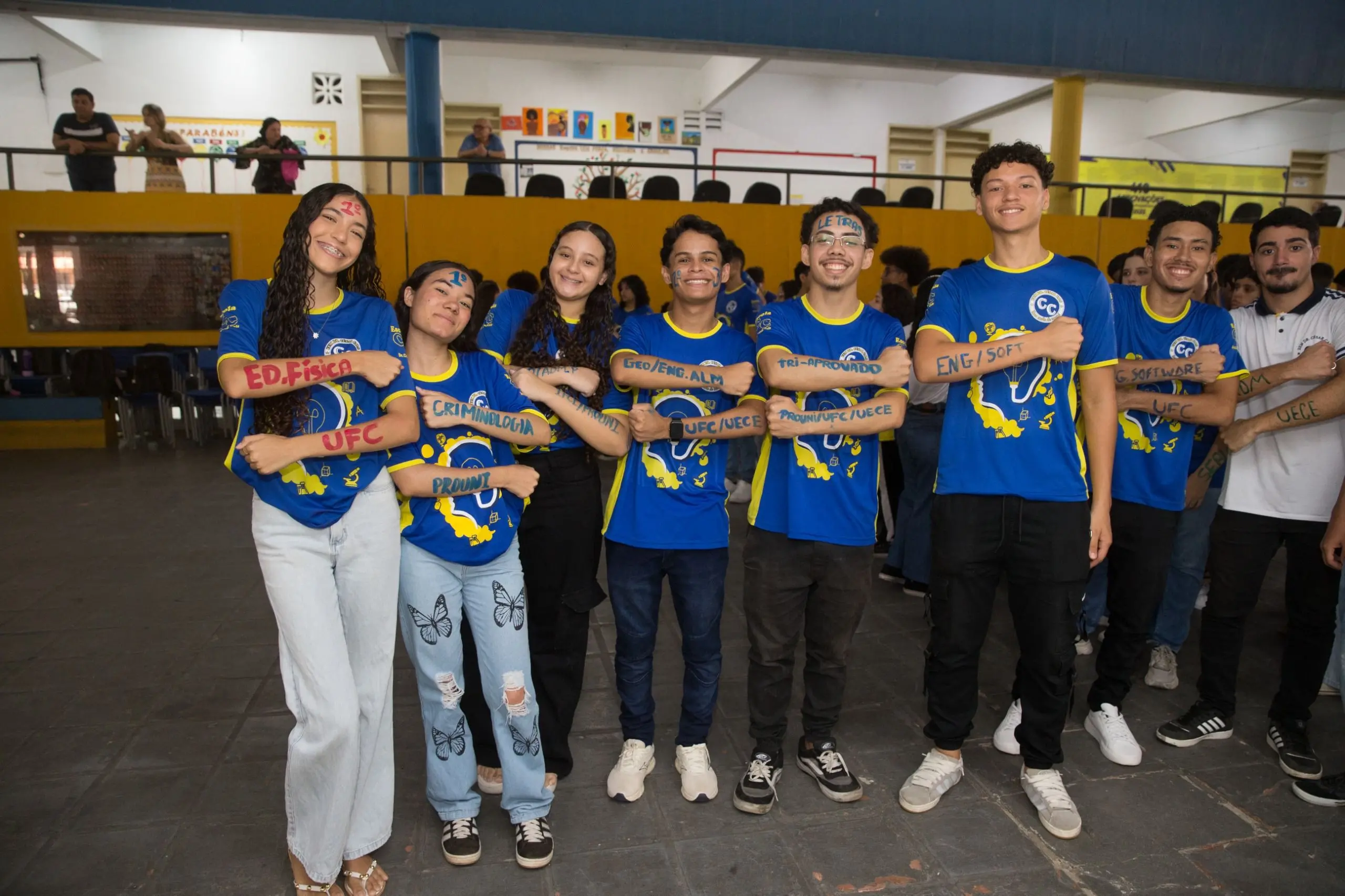 Sete estudantes com fardamento azul celebram a aprovação no vestibular. Eles posam com os braços cruzados, exibindo nomes de cursos e universidades, como UFC e UECE, pintados na pele.
