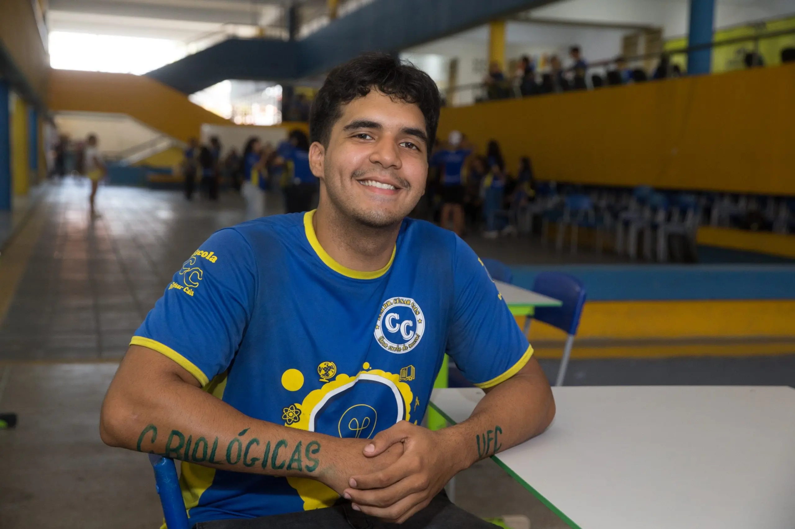 Estudante sorridente com fardamento azul sentado em uma mesa escolar. Nos antebraços, as inscrições Ciências Biológicas e UFC estão pintadas em verde, celebrando sua aprovação acadêmica.