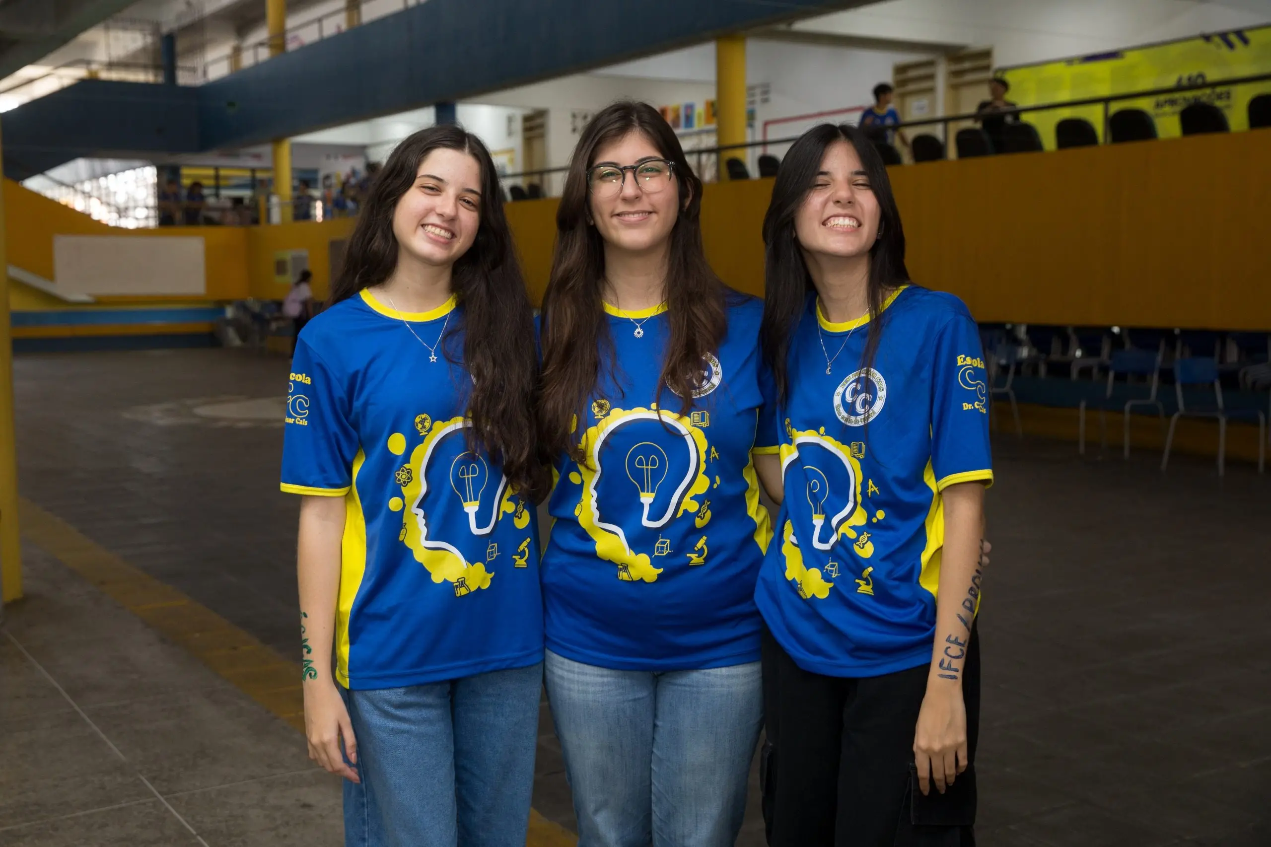 Três jovens mulheres (trigêmeas) sorriem abraçadas em um pátio escolar. Elas vestem camisetas azuis combinando, com detalhes em amarelo e uma estampa de um perfil humano com uma lâmpada dentro da cabeça. A jovem da direita possui a sigla 