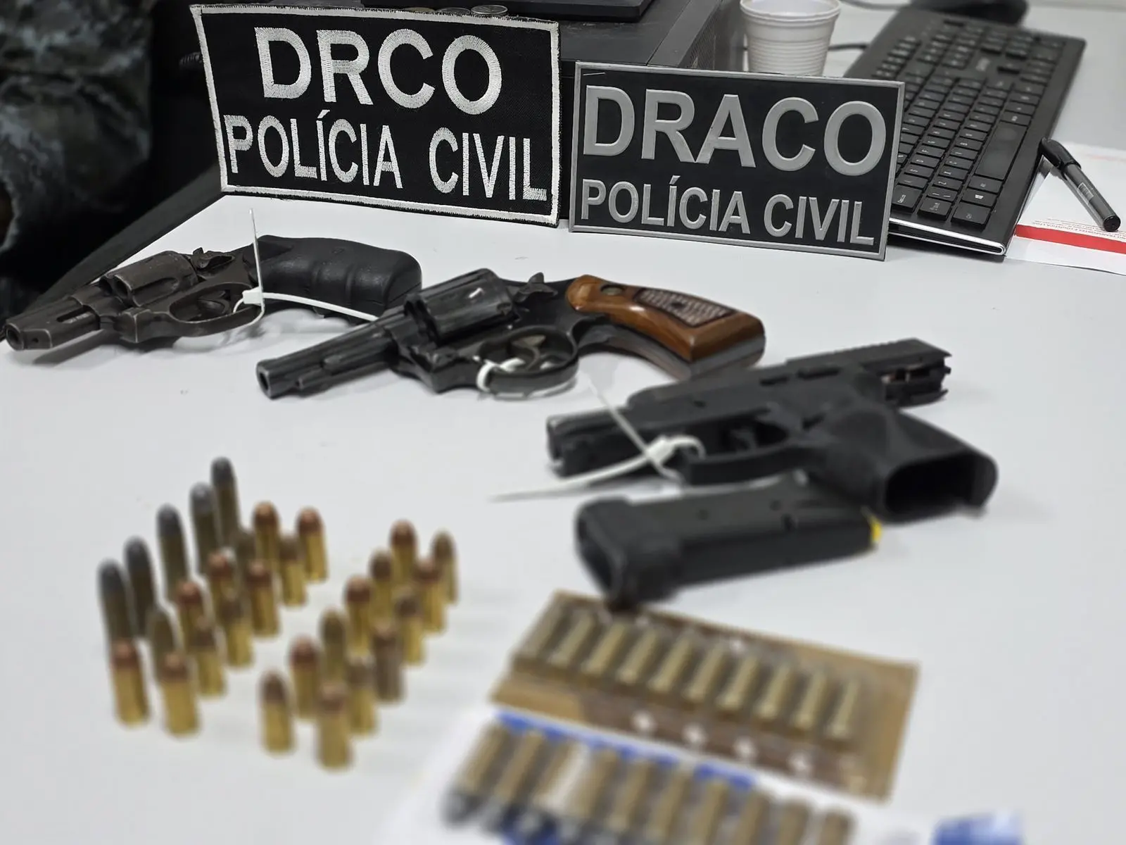 A imagem mostra três armas de fogo e dezenas de munições, apreendidas pela Polícia Civil em uma operação, dispostas em cima de uma mesa, onde há também um teclado de computador, um copo e uma caneta.