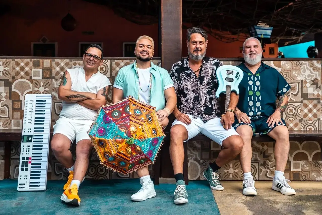 Na imagem, foto promocional da banda Os Transacionais, com quatro integrantes masculinos sentados lado a lado em um banco de madeira, em um ambiente externo com decoração tropical e rústica. Composição e Integrantes: Da esquerda para a direita, o primeiro músico usa óculos, blusa branca sem mangas e tênis amarelos, com um teclado vertical apoiado ao seu lado. O segundo integrante tem cabelo loiro descolorido, veste uma camisa aberta verde-água sobre uma camiseta branca e segura uma sombrinha de frevo colorida e decorada. O terceiro homem tem barba grisalha, veste uma camisa estampada com folhagens e segura uma guitarra azul clara. O quarto músico, à direita, tem barba branca longa e veste um conjunto de camisa e bermuda azul-escuro com estampas geométricas. Cenário: O fundo apresenta uma parede revestida com azulejos de padrões geométricos em tons de bege e marrom. O chão é de cimento queimado em tons de azul e cinza, e o ambiente sugere uma varanda ou quiosque com teto de palha visível na parte superior.