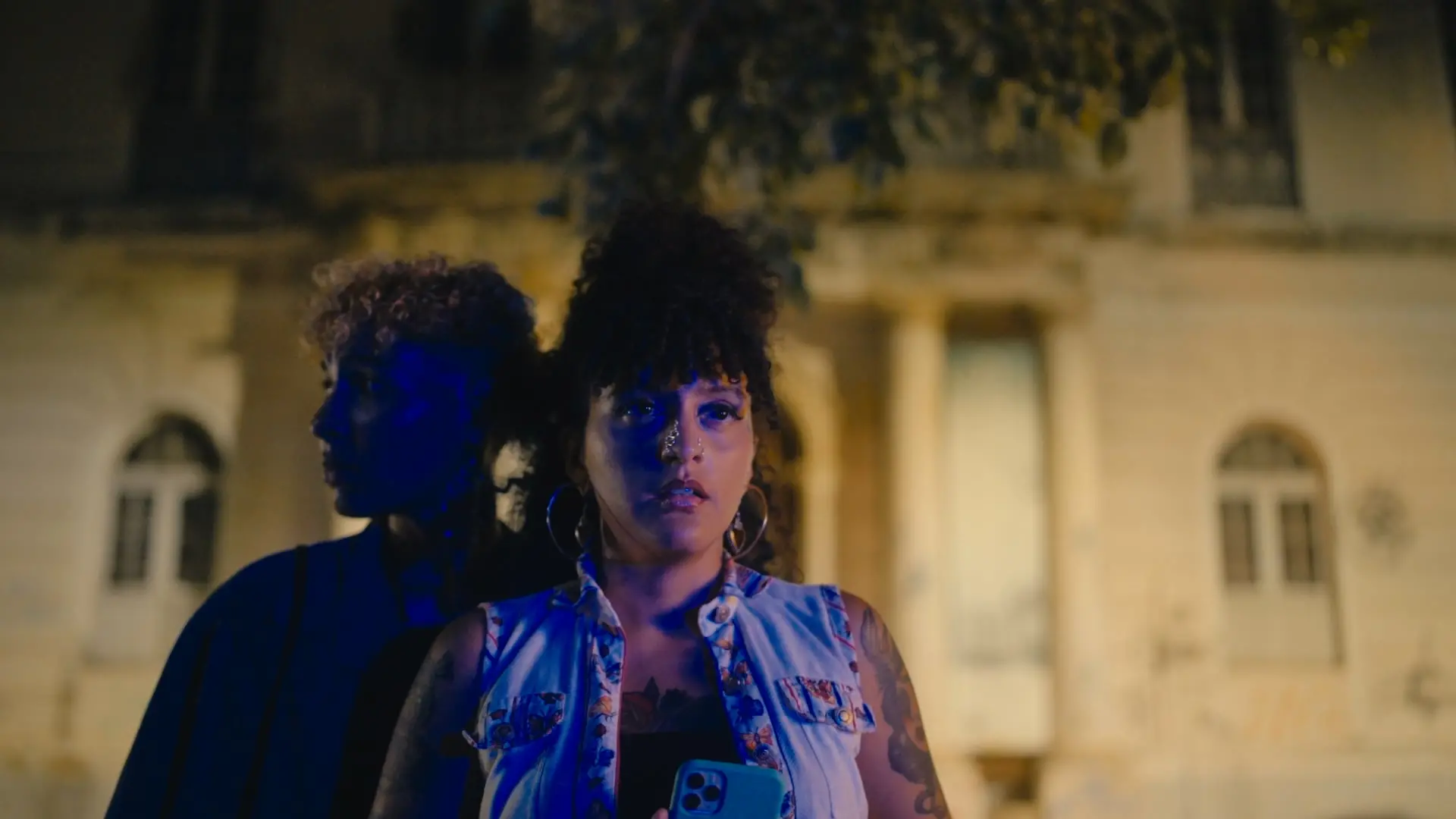 Duas pessoas estão em destaque sob a luz noturna, com uma delas à frente segurando um celular enquanto a outra observa ao fundo, em meio a sombras azuladas. Elas posam diante de um casarão antigo iluminado por luzes amareladas, criando um contraste dramático e urbano que reforça a atmosfera de mistério da cena.