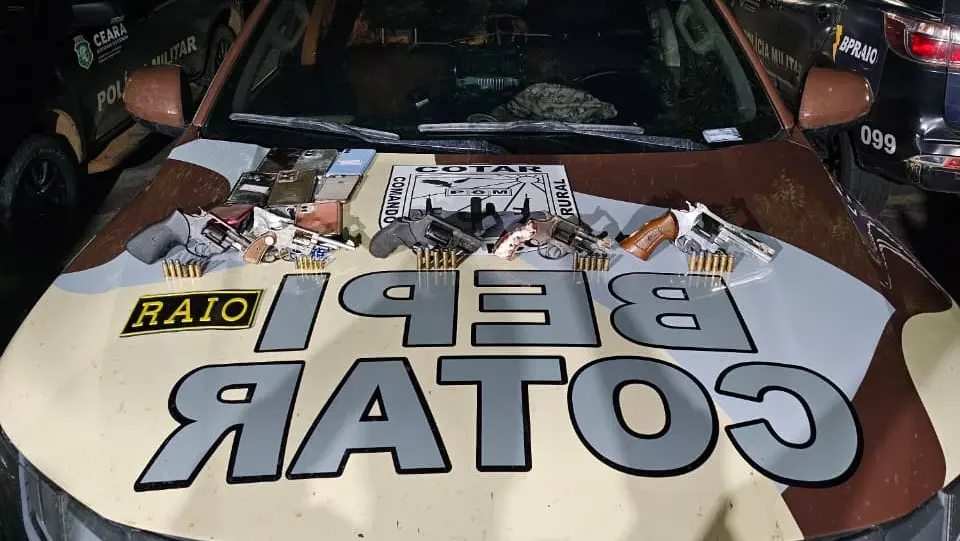 Veículo da Polícia Militar do Ceará com armas, munições, drogas e objetos ilegais espalhados no capô. Cena de operação policial à noite com várias viaturas de polícia ao redor.