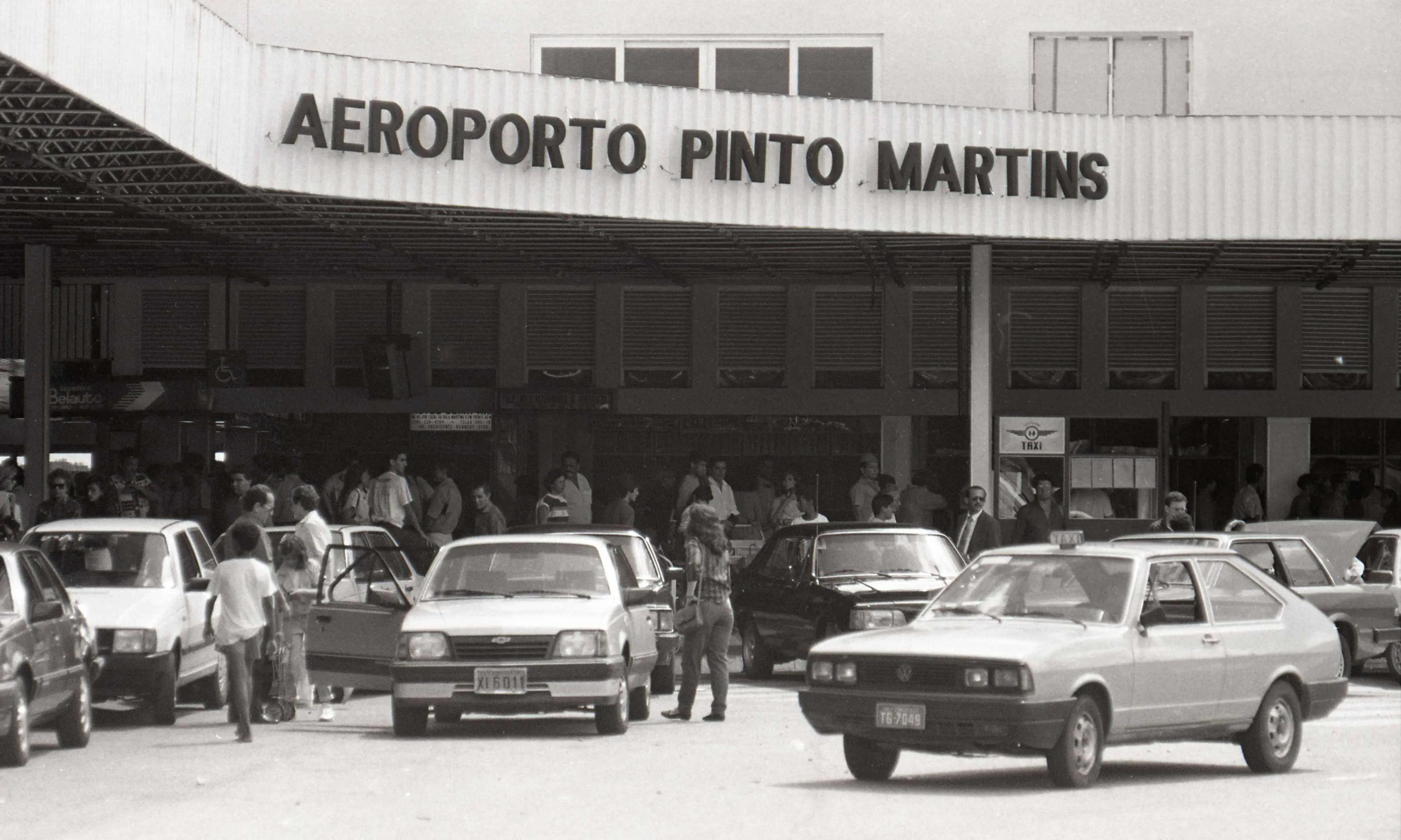 Fachada do antigo Aeroporto Pinto Martins, em Fortaleza, na década de 90. Em frente ao terminal, passageiros e carros de modelos clássicos, como Monza e Passat, movimentam o desembarque.