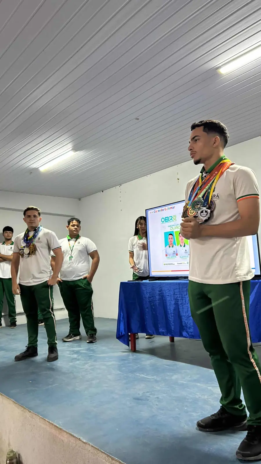 Estudantes com uniformes escolares verdes e brancos e diversas medalhas no peito em um palco. Ao fundo, uma TV exibe o logo da OBR (Olimpíada Brasileira de Robótica) e fotos de premiados.