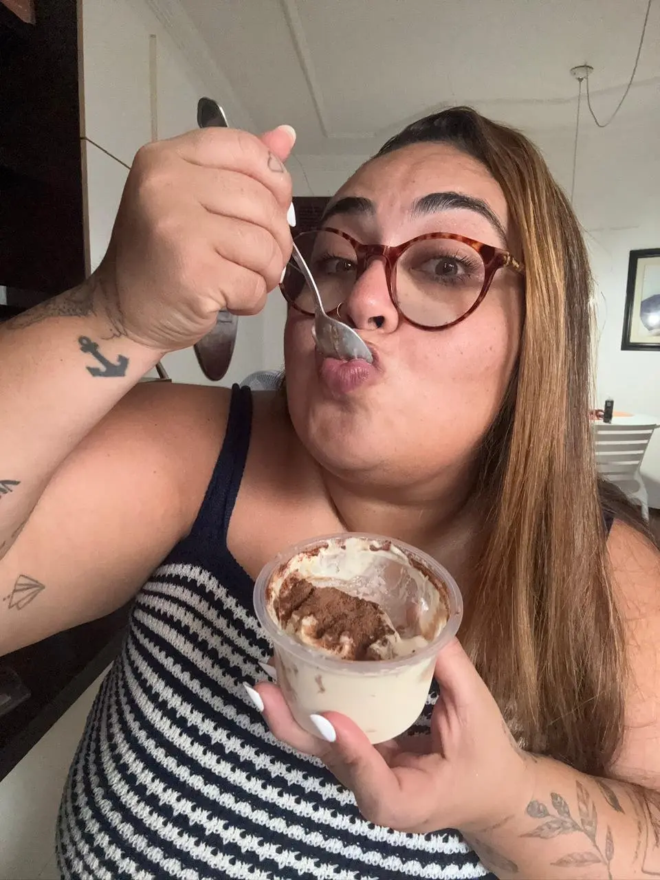 Uma mulher de óculos e cabelos longos segura um pote de sobremesa com pó de cacau, levando uma colher à boca em uma expressão descontraída. Ela usa uma blusa listrada e exibe diversas tatuagens nos braços, em uma composição caseira e iluminada que destaca o prazer de saborear o doce.