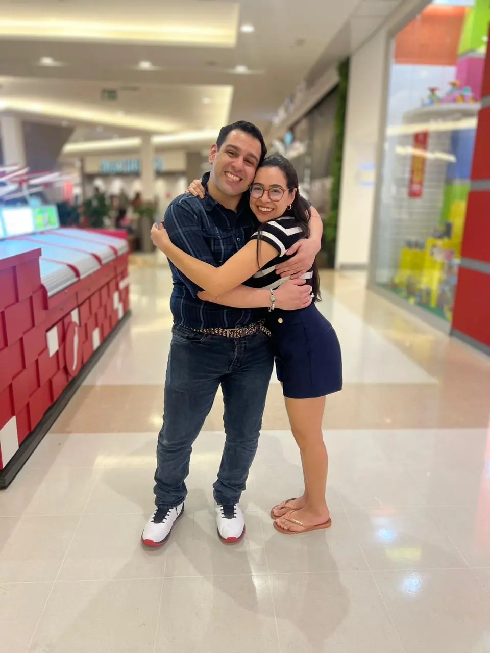 Homem e mulher se abraçando em um shopping center iluminado, próximos a vitrines coloridas e corredores movimentados.