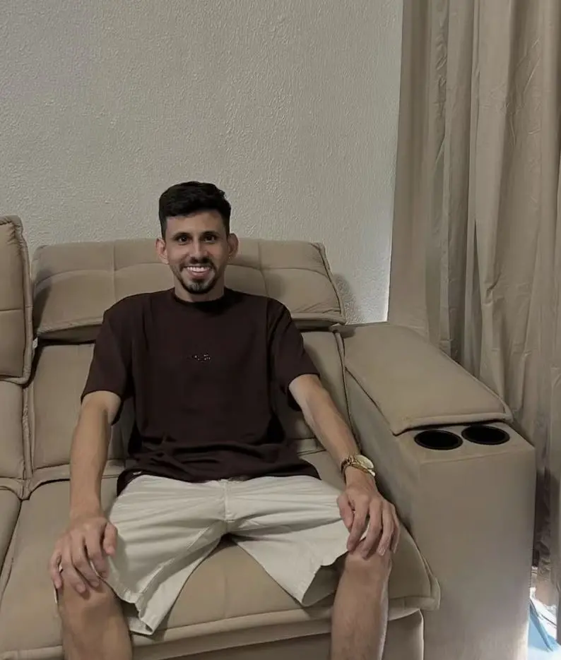 Pessoa sentada em um sofá reclinável bege, vestindo camiseta marrom escura e bermuda clara. As mãos estão apoiadas sobre as pernas e o braço do sofá ao lado possui dois porta-copos embutidos. Há uma cortina longa ao fundo e iluminação suave no ambiente.