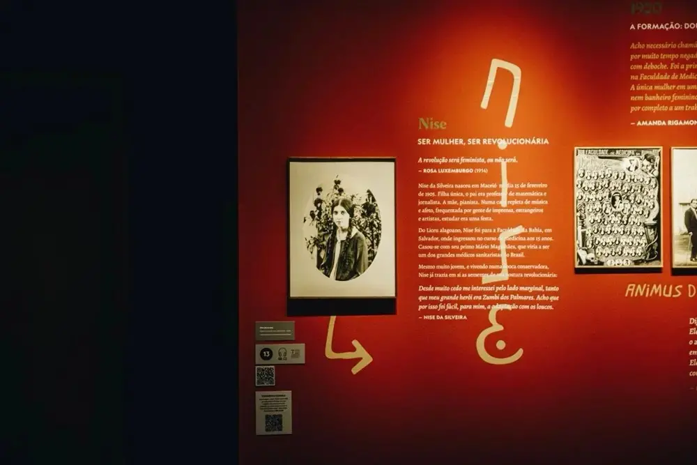 Uma parede vermelha vibrante compõe o cenário da exposição sobre Nise da Silveira, exibindo um retrato em preto e branco da psiquiatra jovem ao lado de textos biográficos e símbolos gráficos em tons de bege. A iluminação focal destaca as informações históricas e as fotografias emolduradas, criando uma atmosfera íntima e informativa na galeria.