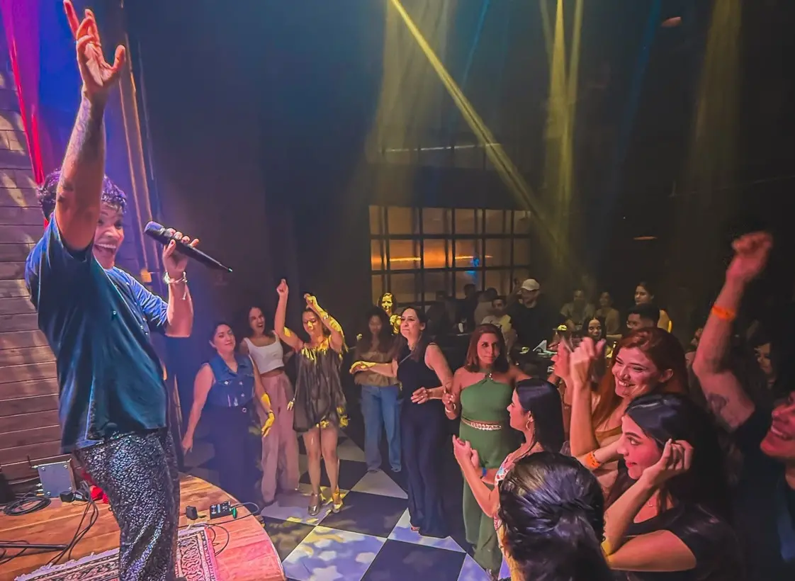 Um cantor se apresenta com entusiasmo em um palco de madeira, vestindo calças brilhantes e erguendo a mão para o público enquanto segura um microfone. Abaixo, uma plateia animada dança e celebra sob feixes de luz amarela que cortam o ambiente escuro do pub, criando uma cena festiva e vibrante.