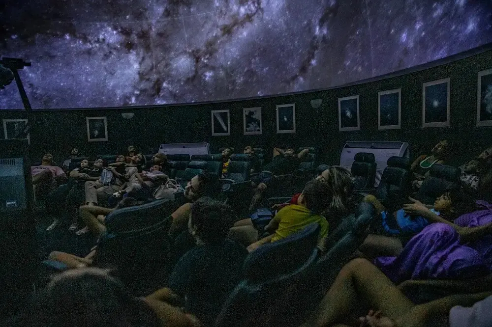 Pessoas acomodadas em poltronas inclinadas observam uma projeção realista que cobre toda a cúpula curva do planetário. O ambiente escuro é pontuado por quadros iluminados nas paredes e pelo brilho azulado das estrelas, capturando um momento de contemplação e descoberta.