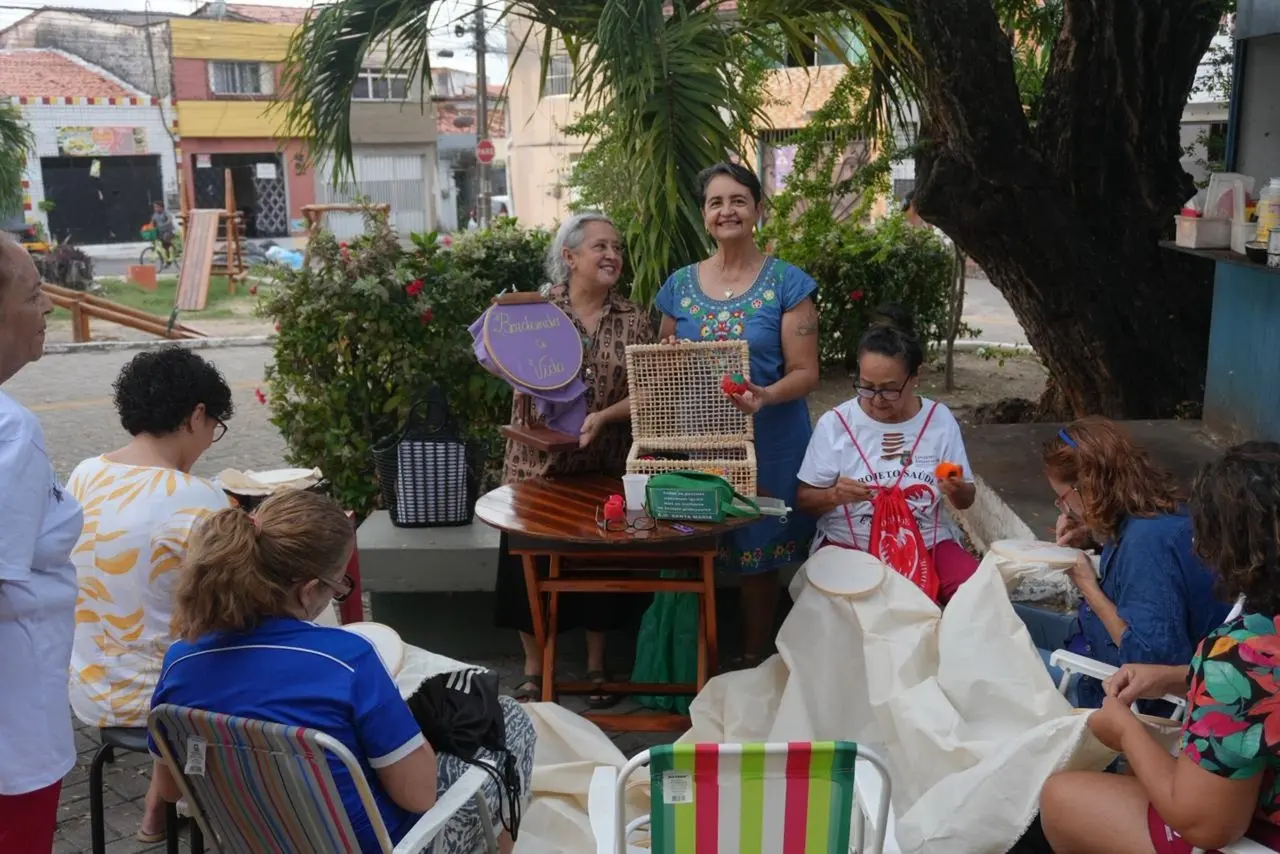 Mulheres participam de uma performance de bordado ao ar livre sob a sombra de uma árvore, exibindo orgulhosamente um bastidor roxo com a frase 