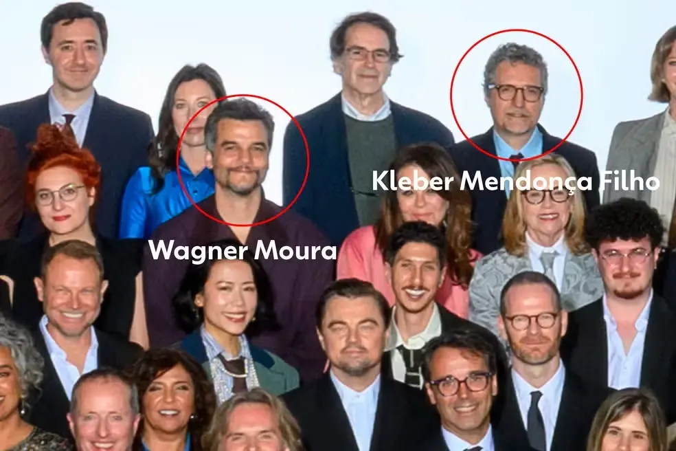 Uma imagem aproximada da foto oficial do Oscar 2026 destaca o ator Wagner Moura e o diretor Kleber Mendonça Filho, ambos circulados em vermelho e identificados por seus nomes em texto branco. Wagner aparece à esquerda com um leve sorriso e camisa escura, enquanto Kleber está à direita usando óculos e paletó azul. Ao redor deles, outras personalidades do cinema, como Leonardo DiCaprio, compõem a multidão de indicados.