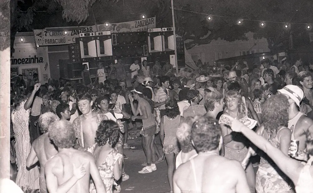 Na imagem, fotografia em preto e branco de uma multidão celebrando o Carnaval de rua em Paracuru, Ceará. A imagem, de ângulo médio e levemente picado, mostra dezenas de foliões, muitos sem camisa ou com roupas leves de verão, dançando em uma área aberta à noite. Ao fundo, destaca-se uma grande estrutura de caixas de som empilhadas (paredão) e faixas com os dizeres 