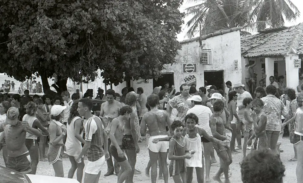 Na imagem, fotografia em preto e branco que retrata uma aglomeração de pessoas durante o Carnaval em Paracuru. A cena se passa em uma rua de terra ou areia, em frente a construções simples de estilo litorâneo. Dezenas de foliões de diversas idades, muitos deles jovens e crianças, aparecem espalhados pela cena. Muitos estão sem camisa, usando trajes de banho ou roupas leves de verão, e diversos aparecem sujos de amido de milho ou farinha, uma tradição comum do Carnaval local.