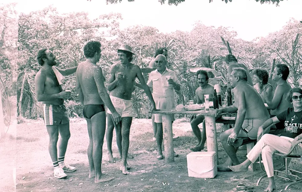 Na imagem, fotografia antiga com tom esverdeado/sépia que registra um momento de confraternização entre amigos em um ambiente ao ar livre, possivelmente em uma área de lazer ou praia. Cerca de dez pessoas estão reunidas em torno de uma mesa rústica de cimento ou pedra. A maioria são homens, alguns sem camisa e usando trajes de banho da época (sungas e shorts curtos), enquanto outros vestem bermudas e camisetas casuais.