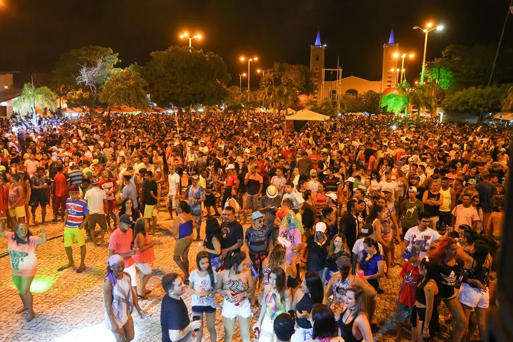 Na imagem, fotografia colorida e noturna que mostra uma multidão densa na Praça de Eventos de Paracuru durante o Carnaval de 2017. A imagem é tirada de um ângulo elevado, oferecendo uma visão panorâmica da celebração. Milhares de pessoas preenchem a praça pavimentada com blocos de concreto claros. A multidão é diversa, vestindo roupas de verão coloridas, como camisetas, regatas e shorts. O clima é de festa intensa, com pessoas dançando e conversando sob a iluminação pública.