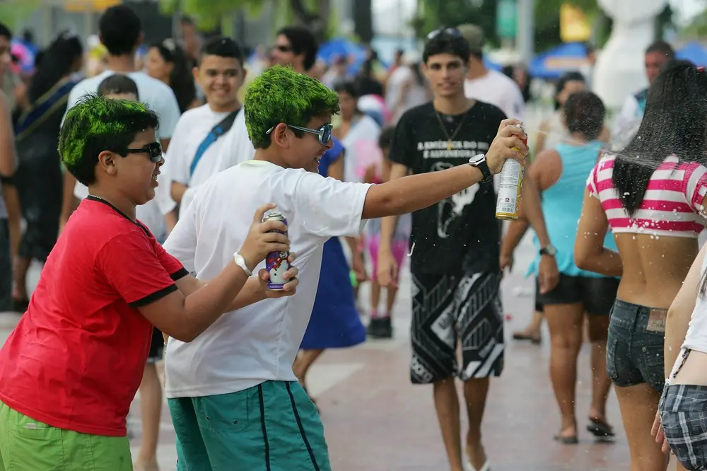 Na imagem, fotografia colorida capturada durante o dia no Carnaval de 2011 em Paracuru. Em destaque, dois meninos com os cabelos pintados de spray verde neon brincam de 