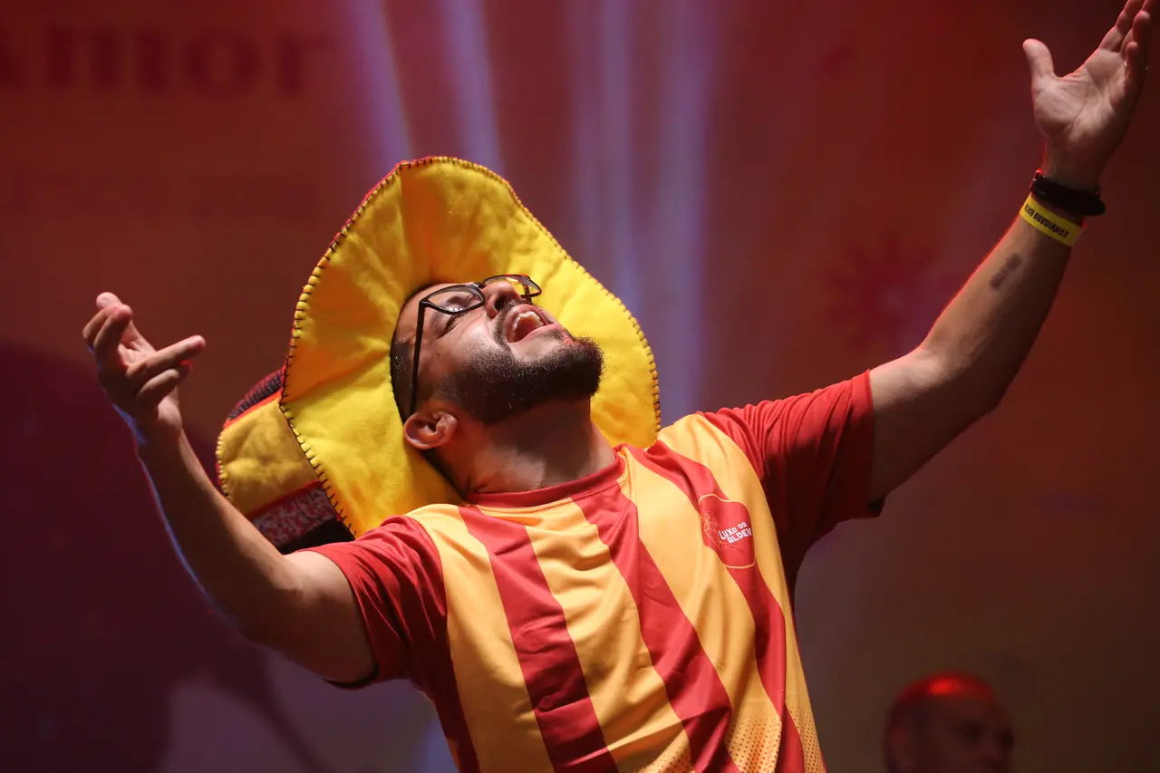 Um integrante do bloco Luxo da Aldeia canta com fervor sob luzes avermelhadas, vestindo uma camisa listrada em amarelo e vermelho e um chapéu estilizado com as mesmas cores. A expressão de entrega e os braços abertos capturam a energia vibrante do pré-carnaval, destacando a paixão do artista no centro da composição.