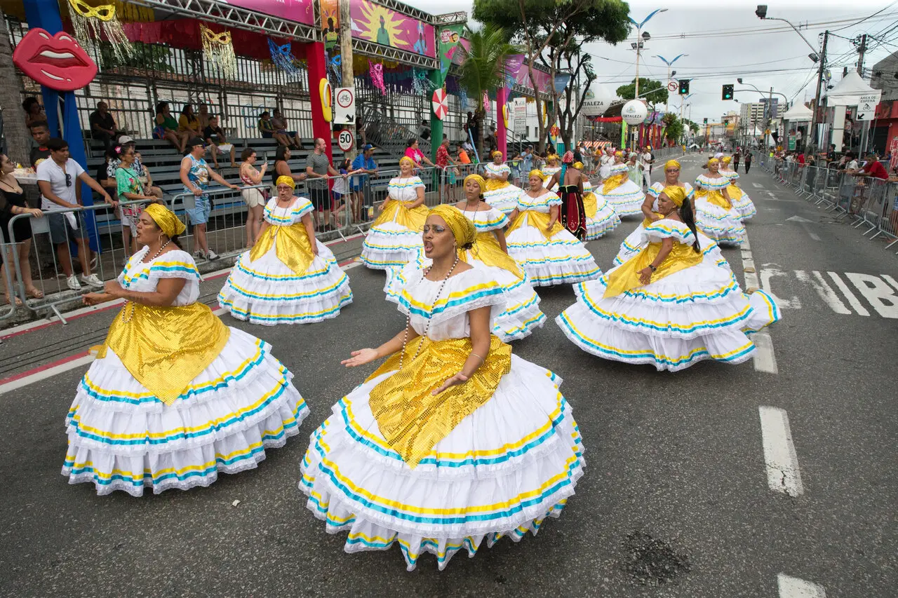 Baianas desfilam pela Avenida Domingos Olímpio vestindo saias rodadas brancas com detalhes em amarelo e azul, enquanto giram em uma coreografia coordenada sob o céu nublado. O cenário urbano de Fortaleza serve de palco para a explosão de tradição e cultura, com o público acompanhando o cortejo pelas arquibancadas laterais.
