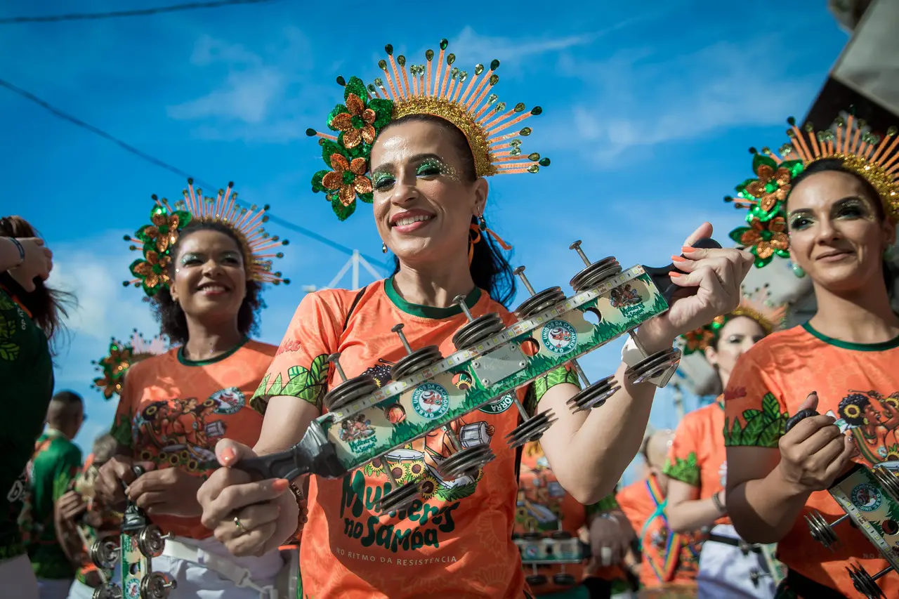 Integrantes da bateria feminina da Unidos da Cachorra sorriem enquanto tocam chocalhos, vestindo camisetas laranjas e adereços de cabeça dourados com flores verdes sob um céu azul claro. A imagem destaca a alegria e a força das mulheres no samba, em uma composição solar repleta de cores e detalhes festivos.