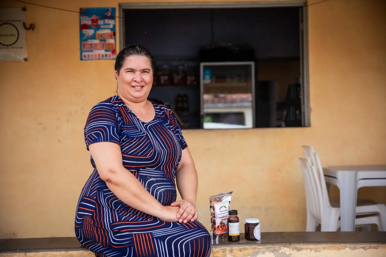 Mulher sentada em frente a pequeno comércio rural exibe produtos à base de caju, como castanhas e doces, apoiados ao seu lado, em ambiente simples e acolhedor.