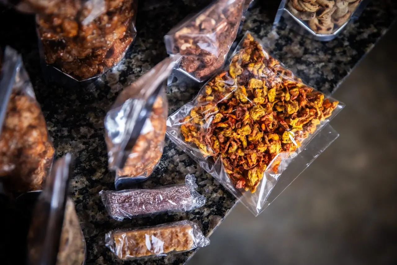 Embalagens plásticas transparentes com produtos derivados do caju, incluindo fibra desidratada usada em preparações veganas, castanhas caramelizadas e doces em barra, dispostas sobre bancada de granito.