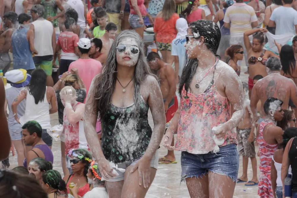 Na imagem, fotografia de uma multidão celebrando o Carnaval de rua em 2015. Em destaque no centro, duas jovens caminham cobertas de pó branco (maisena ou talco) da cabeça aos pés. A jovem à esquerda usa óculos escuros espelhados e uma regata preta com estampas circulares; a jovem à direita usa uma regata floral rosa e shorts jeans. Ao redor delas, dezenas de foliões também aparecem sujos de pó, alguns com cabelos coloridos de tinta verde e rosa. O ambiente é ensolarado e festivo, transmitindo uma atmosfera de celebração popular.