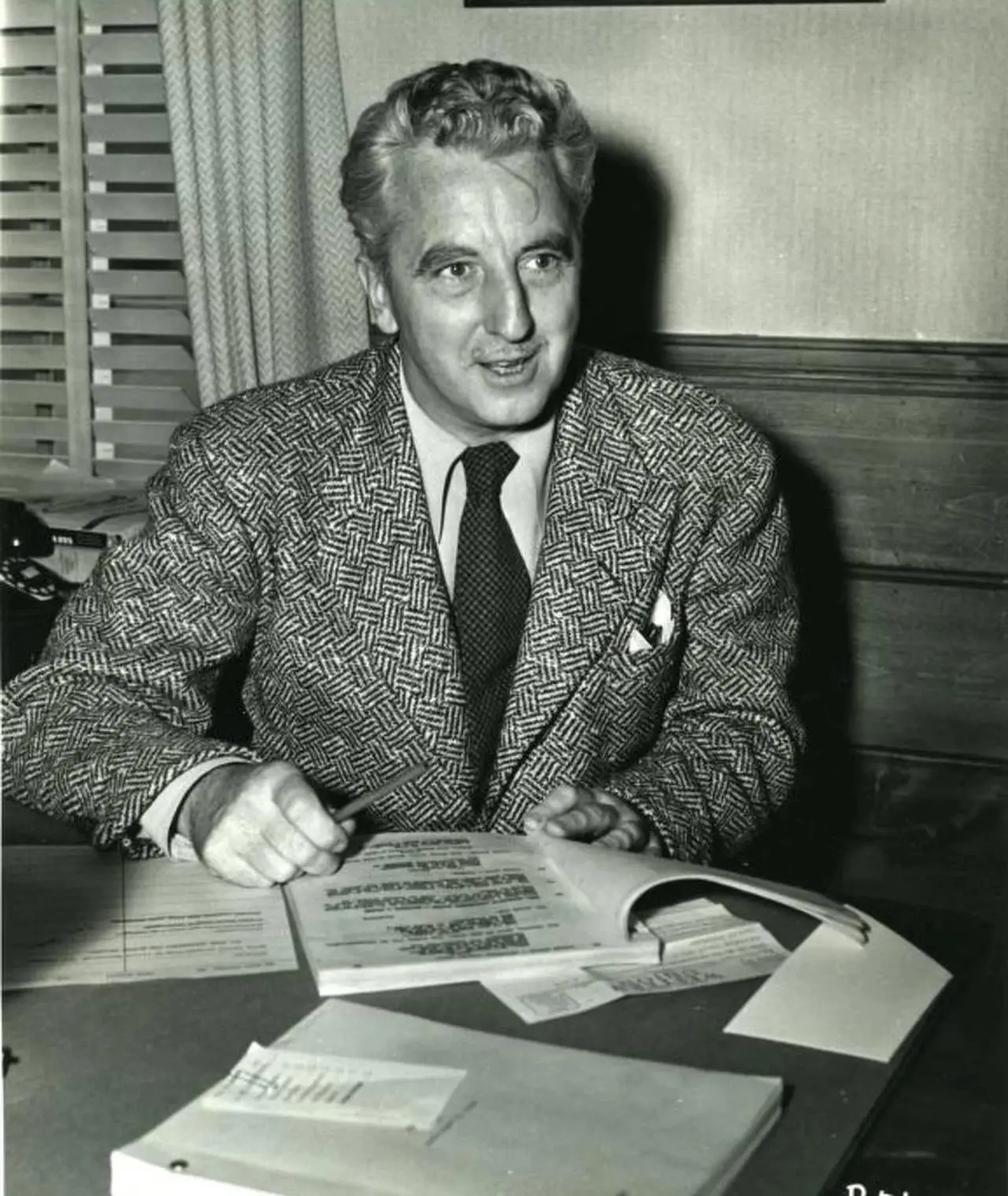 Homem de terno com cabelo ondulado sentado em uma mesa com documentos, sorrindo durante uma conversa, ambiente de escritório com cortinas ao fundo.