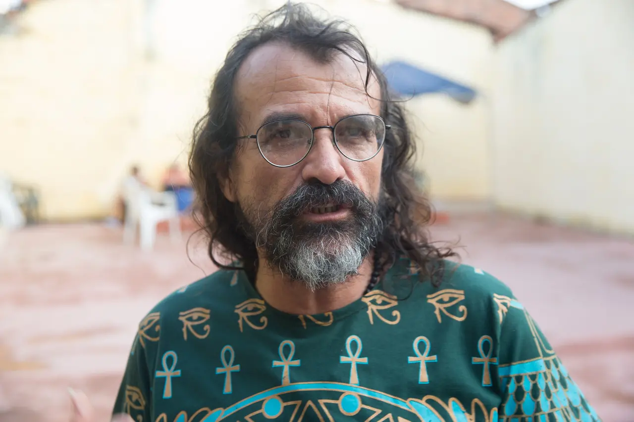 Pingo de Fortaleza posa para um retrato em close, usando óculos redondos e uma camiseta verde estampada com símbolos egípcios como o Olho de Hórus e o Ankh. O artista possui cabelos longos ondulados e barba grisalha, apresentando uma expressão serena em um ambiente externo levemente desfocado.
