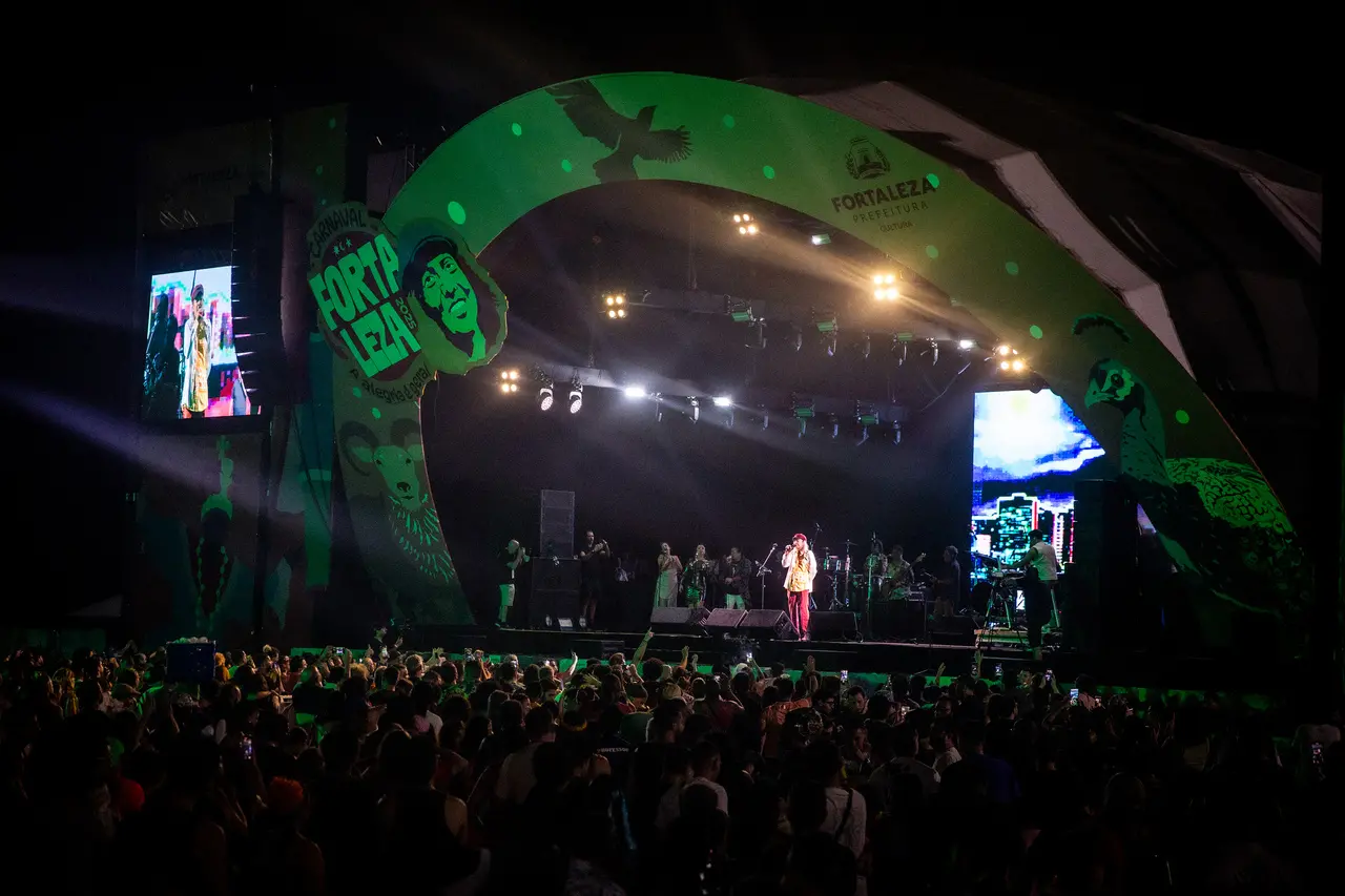 O cantor Ednardo se apresenta em um palco iluminado sob um grande arco verde decorado, enquanto uma multidão entusiasmada acompanha o show durante a noite. O cenário destaca painéis de LED e uma iluminação vibrante que compõe a atmosfera festiva do Carnaval de Fortaleza no Aterrinho da Praia de Iracema.