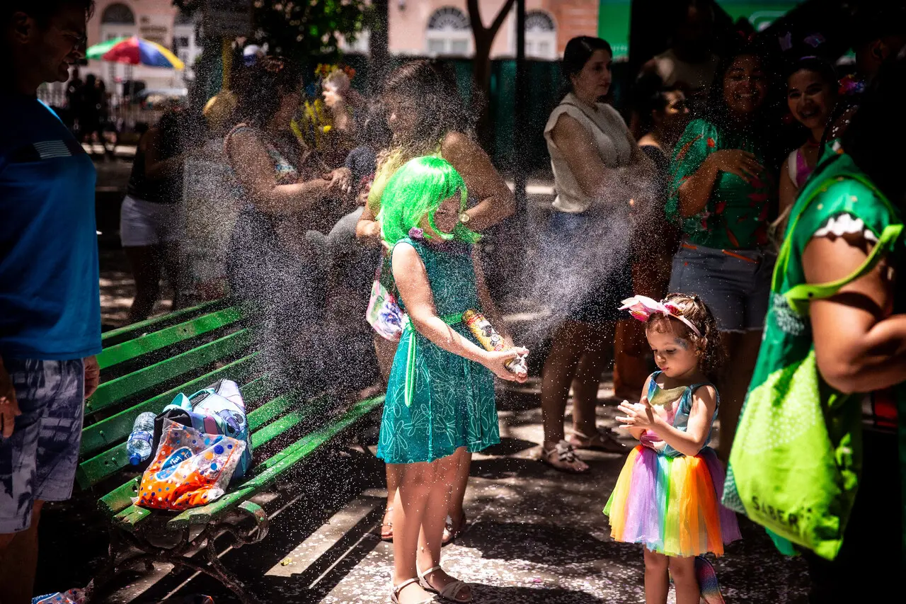 Uma criança vestida com peruca verde e vestido brilhante diverte-se lançando spray de espuma no Passeio Público, enquanto outra menina com saia colorida de tule observa a brincadeira. Ao redor, adultos acompanham a cena em meio às sombras das árvores, capturando um momento lúdico e refrescante do Carnaval infantil em Fortaleza.