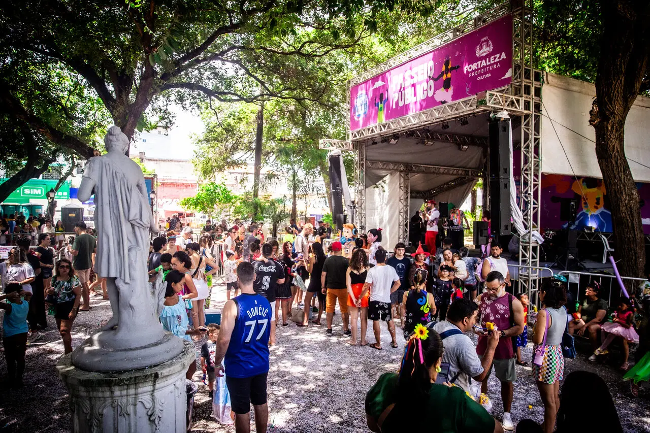 Registro de movimentação festiva no Passeio Público, onde famílias e foliões se reúnem em frente a um palco sob a sombra das árvores. O cenário é marcado por um pórtico rosa com a inscrição 