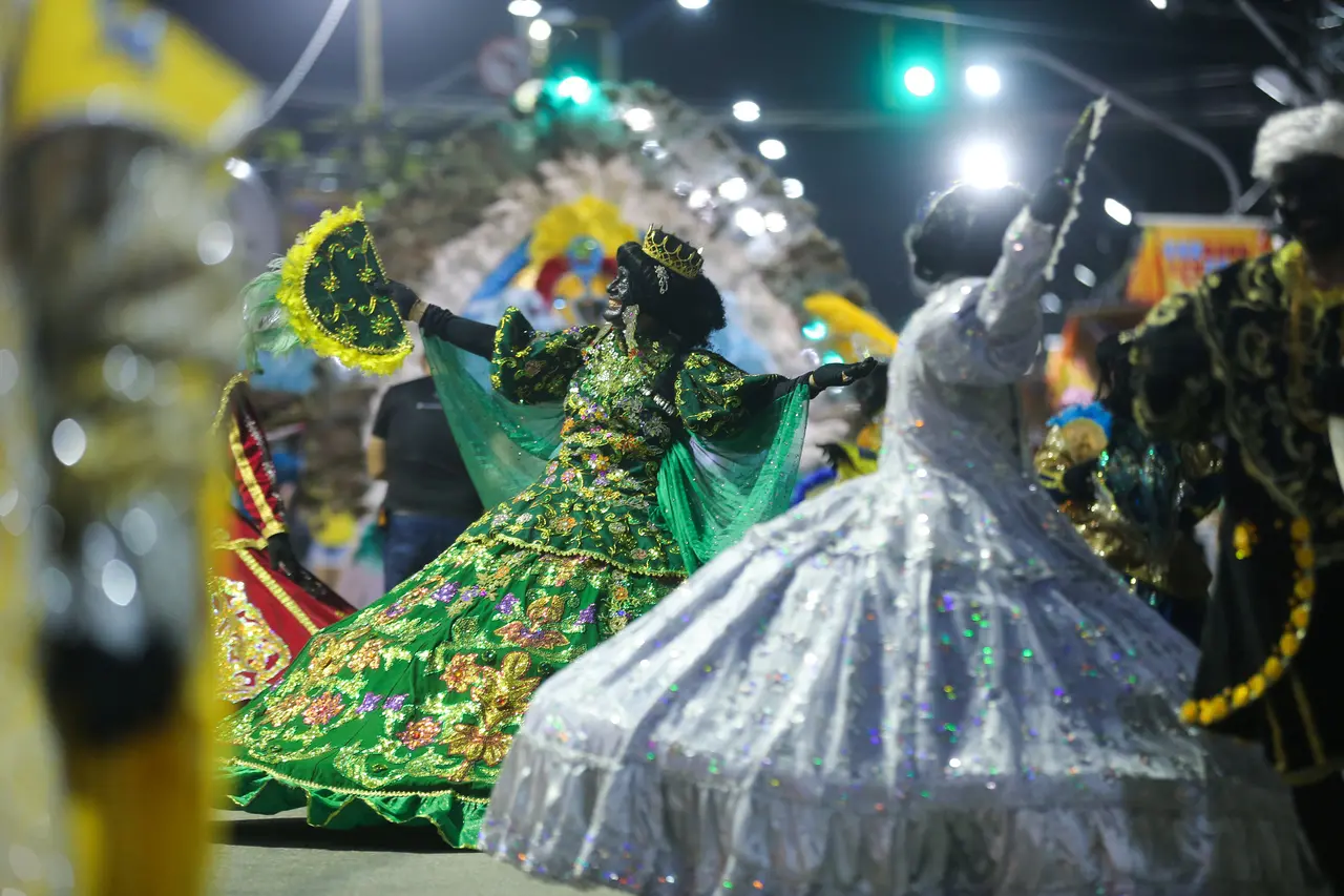 Integrantes de um Maracatu desfilam na Avenida Domingos Olímpio, com destaque para uma figura central em um suntuoso vestido verde bordado com flores douradas e uma pequena coroa. Ao seu lado, outra baliza em um vestido branco volumoso gira sob as luzes noturnas, compondo uma cena de celebração tradicional com cores vibrantes e movimento.
