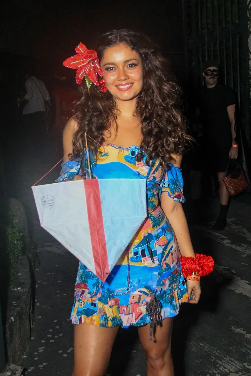 Pessoa usando um vestido colorido estampado com casas e paisagens, segurando um grande objeto em forma de pipa nas cores azul, branco e vermelho. Ela tem cabelos longos e cacheados e usa um adereço vermelho chamativo preso ao cabelo. Em um dos pulsos, há um acessório vermelho volumoso. A cena ocorre à noite, em uma área externa pouco iluminada.