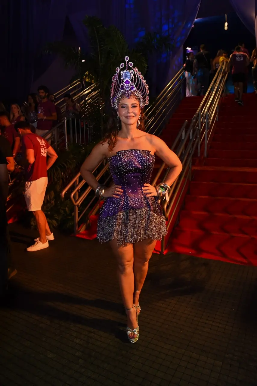 Mulher vestindo um look carnavalesco composto por um vestido curto tomara que caia em tons de roxo e prata, coberto por brilhos e pedrarias. Ela utiliza um grande adereço de cabeça ornamentado com detalhes roxos, prateados e pedras reluzentes. Está parada no centro de um corredor com tapete vermelho e corrimões metálicos ao fundo, em um ambiente iluminado e decorado para um evento festivo. Pessoas circulam ao redor, reforçando o clima de celebração.