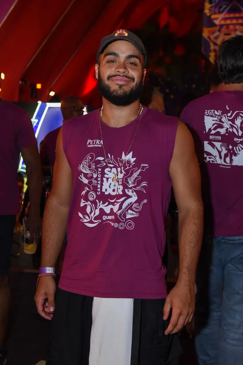 Homem usando uma camiseta regata roxa estampada com ilustrações e logos de camarote de Carnaval. Ele está em pé em um ambiente festivo e iluminado, com outras pessoas ao fundo vestindo roupas semelhantes. Usa um boné, colar e pulseira, e posa diretamente para a foto em meio à movimentação do evento.